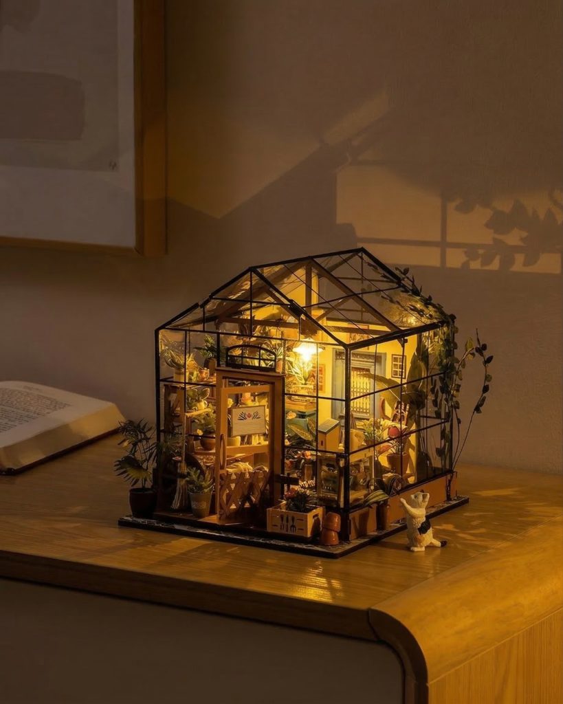 A finished miniature greenhouse kit glowing with warm interior light, displayed on a wooden desk alongside a book with plant shadows on the wall.